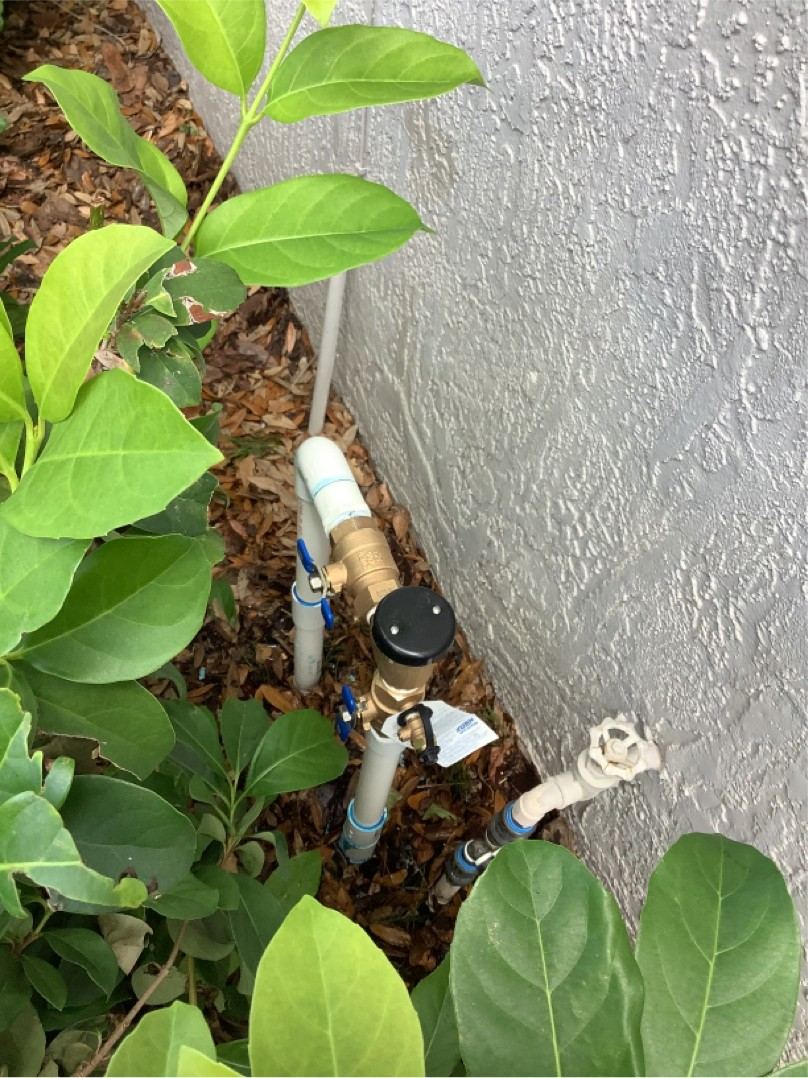 Brass backflow preventer installed on PVC pipework against a house wall with a test tag attached