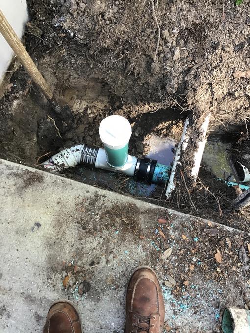 Newly installed PVC sewer clean out with a green cap set in a trench beside a concrete walkway