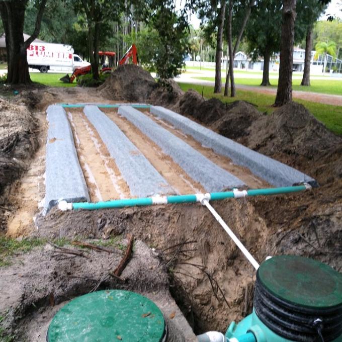 Drain field laterals wrapped in geotextile fabric with green distribution piping, excavator and work truck behind