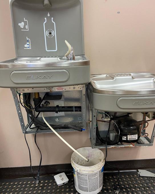 Drinking fountain covers removed to expose the chiller units and a drain line running into a bucket