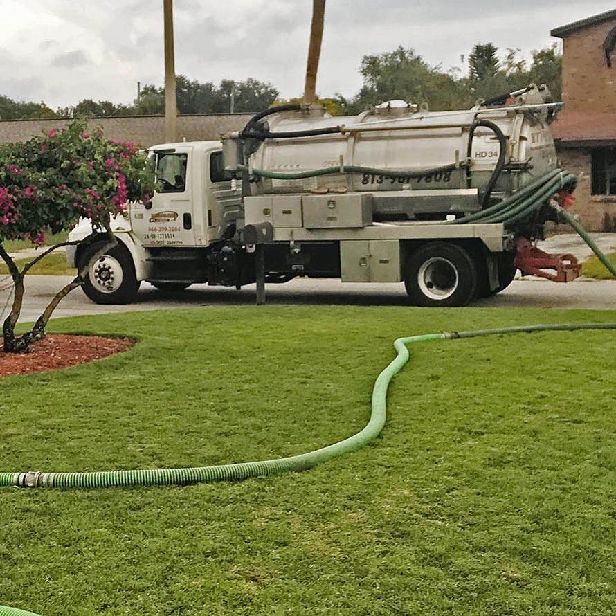 Tampa Bay Plumbers septic pump truck parked at the curb with suction hoses run across a front lawn to the tank
