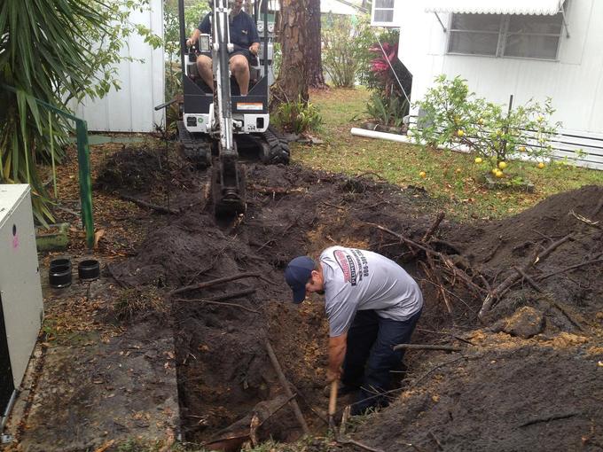 Mini excavator and a Tampa Bay Plumbers technician working in an open trench beside a house during a sewer line replacement