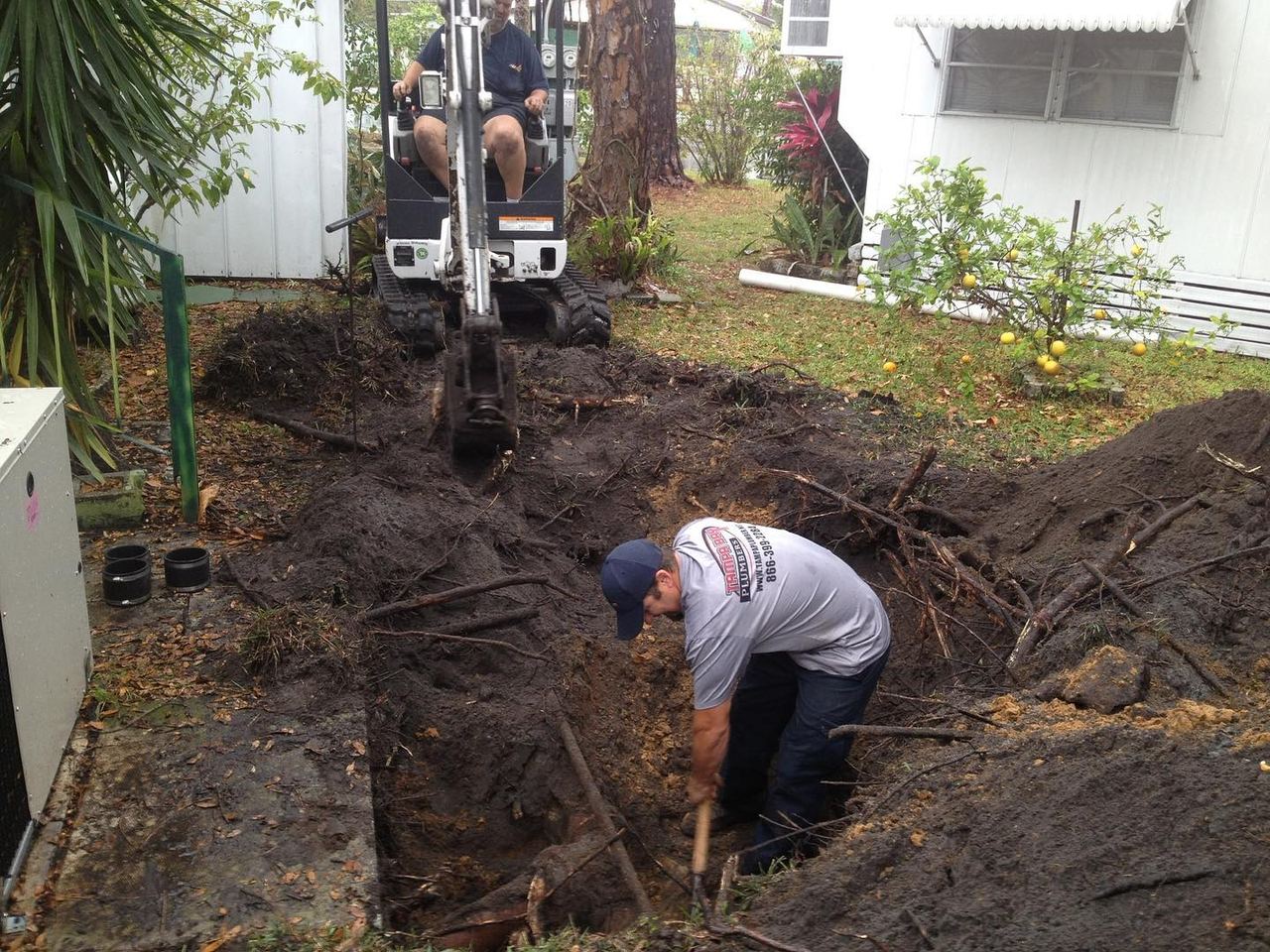 Mini excavator and a Tampa Bay Plumbers technician working in an open trench beside a house during a sewer line replacement