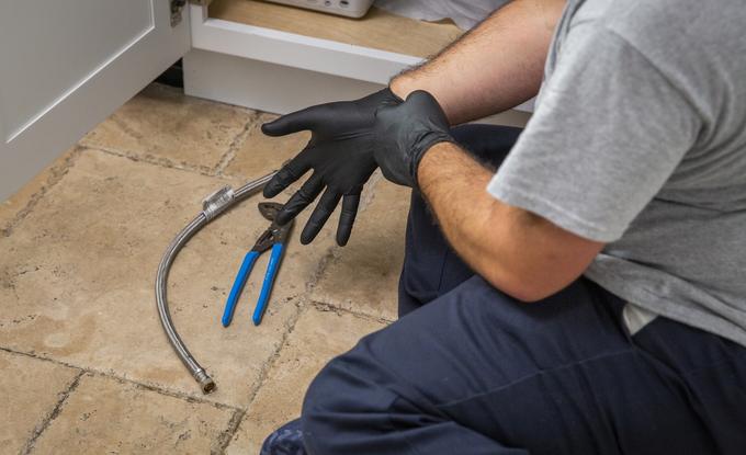 Technician gloving up beside a braided supply line and pliers under a kitchen cabinet