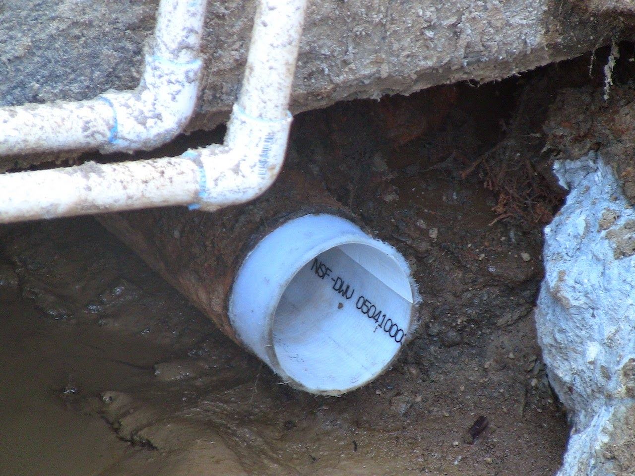 New white PVC spliced into an old corroded drain line in a wet excavation beneath a concrete slab