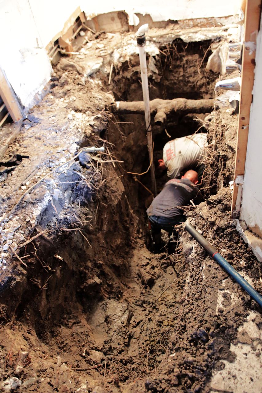 Floor opened inside a house down to a deep trench full of tree roots with a technician working on the drain line below