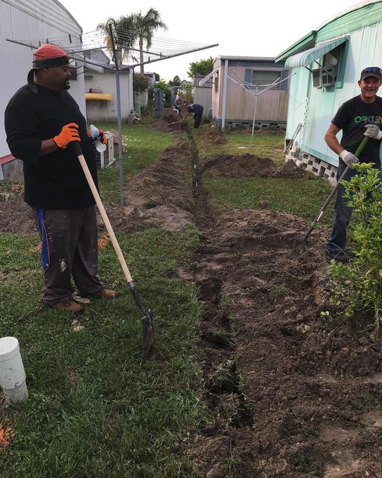 Two Tampa Bay Plumbers crew members hand digging a trench between manufactured homes for a new main water line